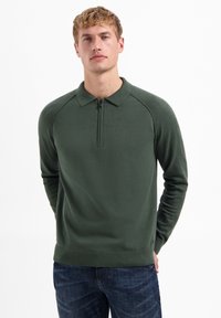 Donkergroene pullover met een kwart-rits kraag, geribbelde zoom en getextureerde schoudernaden. Draagt met donkerdenim jeans.