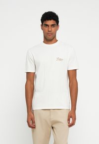 Camiseta de algodón blanca con mangas cortas, escote redondo y pequeño logo bordado en marrón en el lado izquierdo del pecho. Diseño simple y limpio.