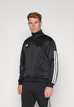 Sort atletisk jakke med høj krave, lynlås fortil og tre hvide striber på ærmerne. Lavet af glat, letvægtsstof.