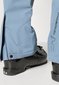 Világoskék sínadrág, a térdnél texturált, geometrikus mintával, cipzárakkal és kiegészítőkkel. Fekete síbakancsok láthatók alatta.