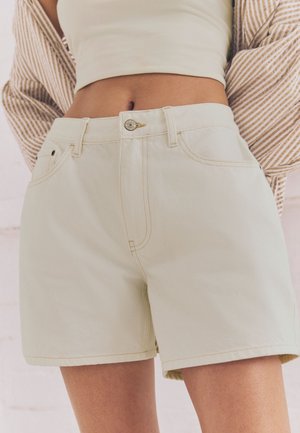 Mujer con shorts vaqueros de tiro alto en beige claro, top corto y chaqueta a rayas con las manos detrás de la espalda.