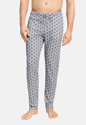 Uomo con pantaloni del pigiama grigio chiaro con un piccolo motivo geometrico rosso e nero, in piedi a piedi nudi su uno sfondo semplice.