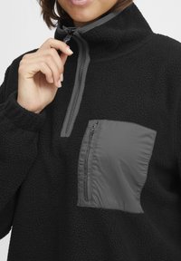 Must musta pullover, millel on veerandi lukuga kaelus, kontrastne hall lukuga rinna tasku, tekstuuriga kangas ja hallid aktsendid.