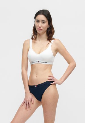 Γυναίκα που φοράει λευκό bralette Tommy Hilfiger και σκούρο μπλε εσώρουχα, στέκεται με το ένα χέρι στη μέση της μπροστά από μονόχρωμο φόντο.