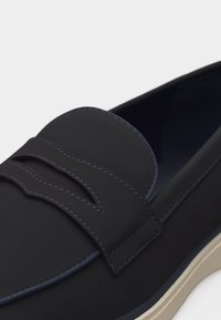 Mocassin bleu marine foncé à enfiler avec une bride sur le dessus, semelle beige clair contrastante et surpiqûres apparentes.