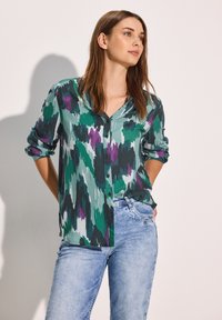 Camicia con bottoni realizzata in un tessuto texturizzato con un motivo astratto nei colori verde, viola e nero. Maniche lunghe arrotolate, abbinata a jeans azzurri chiari.