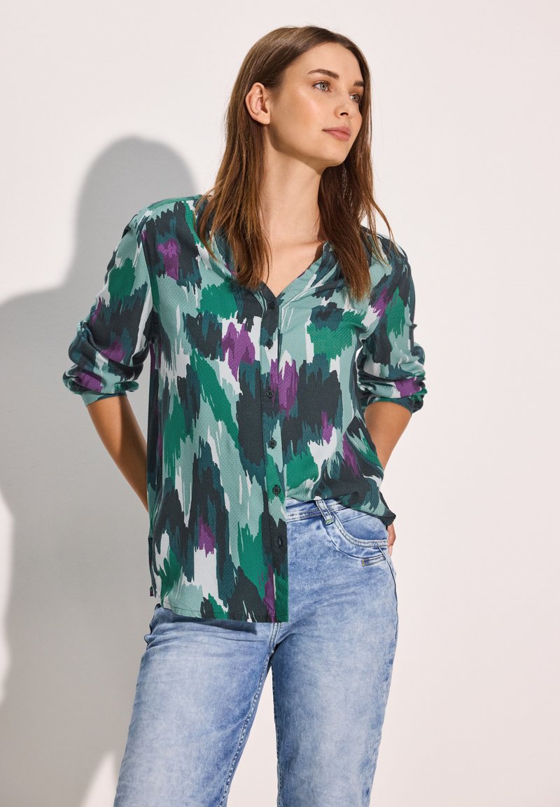 Camicia con bottoni realizzata in un tessuto texturizzato con un motivo astratto nei colori verde, viola e nero. Maniche lunghe arrotolate, abbinata a jeans azzurri chiari.