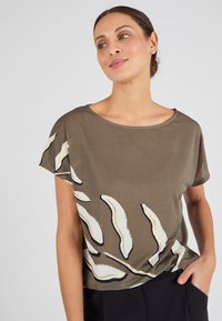 Modelo con una camisa suelta de color verde olivo con patrones abstractos de hojas en blanco y negro, combinada con pantalones negros, sonriendo suavemente contra un fondo claro.