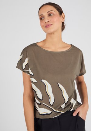 Modelo con una camisa suelta de color verde olivo con patrones abstractos de hojas en blanco y negro, combinada con pantalones negros, sonriendo suavemente contra un fondo claro.