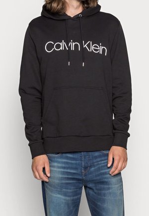 Person iført sort Calvin Klein-hoodie og blå jeans, stående med hænder afslappet ned langs siderne mod en ensfarvet baggrund.