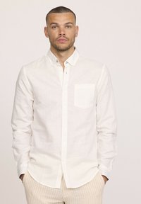 Chemise boutonnée en lin blanc avec une poche, col décontracté et manches longues. Texture subtile, associée à un pantalon beige à rayures.