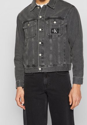 Mężczyzna w wyblakłej czarnej kurtce dżinsowej z zapinanymi na guziki kieszeniami na piersi, z naszywką z logo CK Jeans, w połączeniu z czarnymi dżinsami.