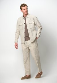 Giacca in lino beige chiaro con due tasche sul petto, abbinata a un maglione marrone e pantaloni beige coordinati, completata da scarpe slip-on marroni.