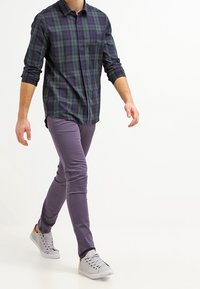 Rutig skjorta i mörkblått och grönt, kombinerad med lila slim-fit byxor och grå sneakers med enkla snören och en texturerad design.