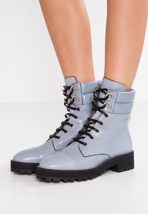 Bottes montantes lacées gris clair brillantes avec semelles épaisses noires portées sur des jambes nues sur un fond blanc uni.