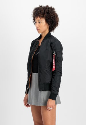 Veste bomber noire avec des bords côtelés, doublure orange et poches latérales. Présente une étiquette rouge sur la manche et une texture lisse et brillante.