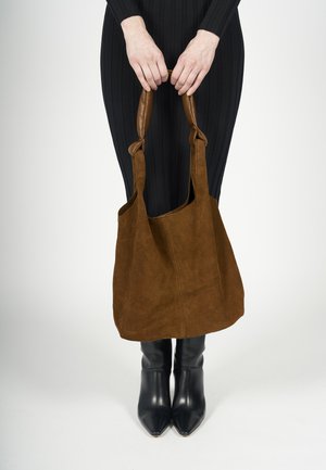 Brun ruskind tote taske med to læderhåndtag, struktureret form og blød tekstur, holdt af en person iført et sort ribbet outfit og ankelstøvler.