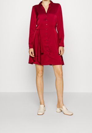 Femme portant une robe rouge à manches longues avec des boutons, un ourlet asymétrique et des chaussures crème à talons blocs bas à enfiler.