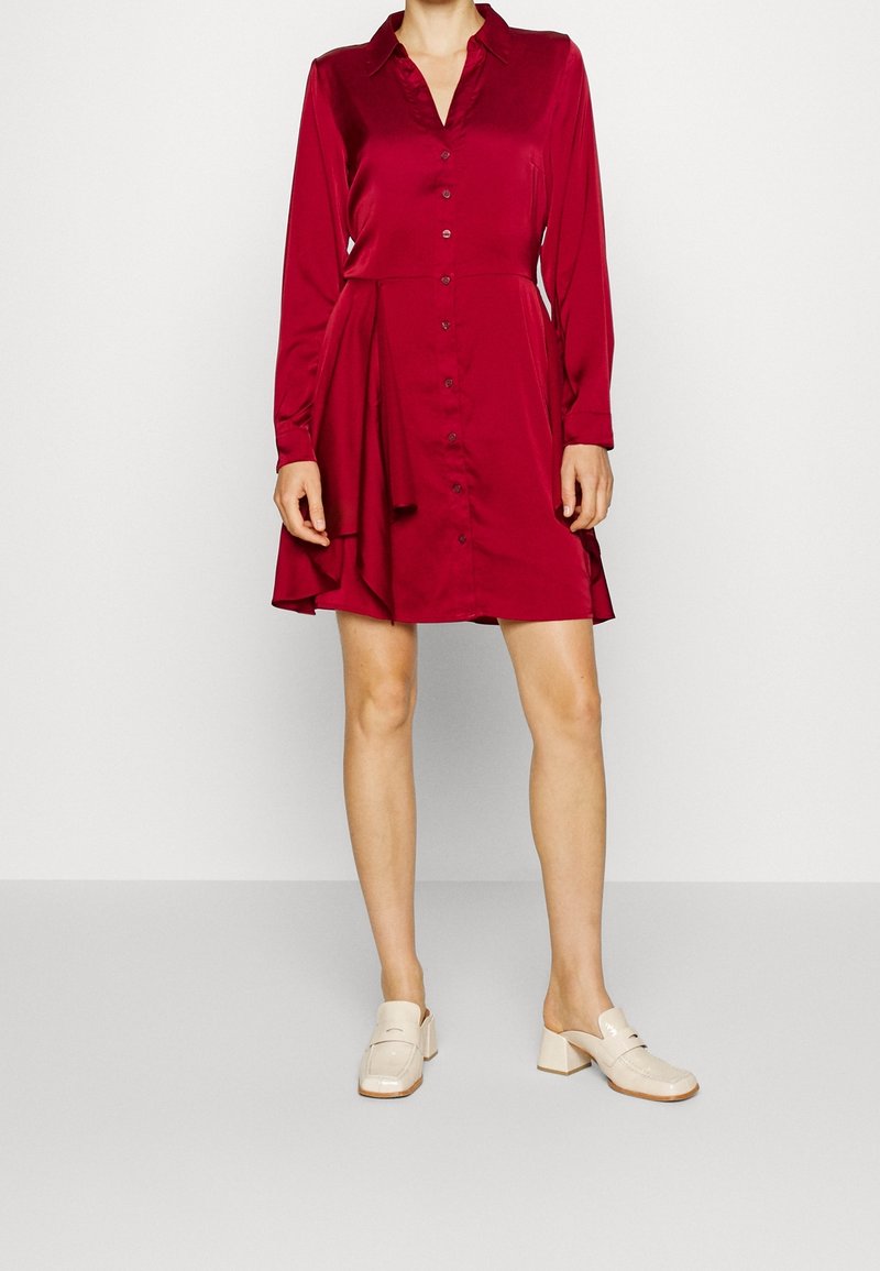 Femme portant une robe rouge à manches longues avec des boutons, un ourlet asymétrique et des chaussures crème à talons blocs bas à enfiler.