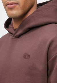 Κοντινή λήψη ενός ατόμου που φοράει ένα μπορντό φούτερ με μία μικρή κεντημένη λογότυπο στο στήθος, που δείχνει το κάτω μέρος του προσώπου και τον λαιμό.