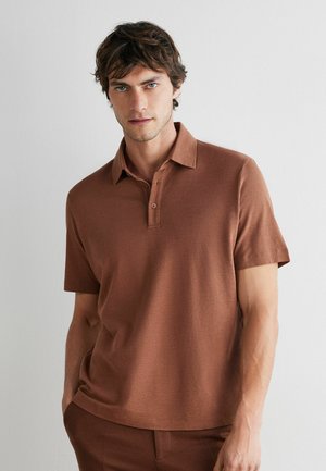 Mężczyzna o brązowych włosach, ubrany w brązową krótką koszulkę polo i pasujące spodnie, stojący na tle gładkiej białej ściany.