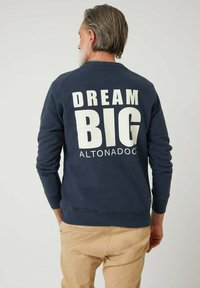 Hombre con cabello y barba canosos, vistiendo un sudadera azul marina con el texto "DREAM BIG ALTONADOCK" en la espalda, pantalones beige, de pie contra un fondo blanco.