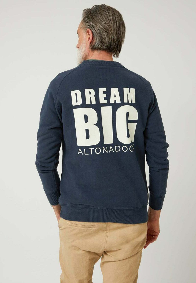 Hombre con cabello y barba canosos, vistiendo un sudadera azul marina con el texto "DREAM BIG ALTONADOCK" en la espalda, pantalones beige, de pie contra un fondo blanco.