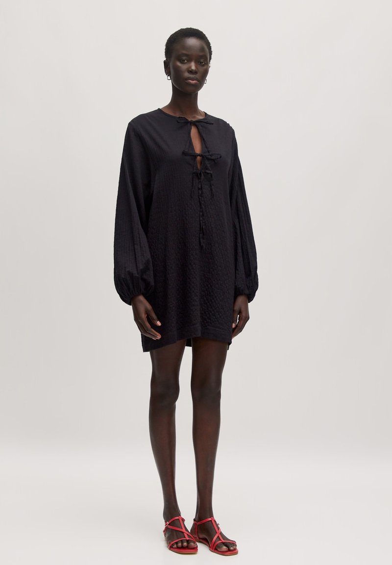 Schwarzes, strukturiertes Tunika-Kleid mit einer Bindeschließe, langen Puffärmeln und einer lockeren Passform. Kombiniert mit roten Sandalen mit dünnen Riemen.