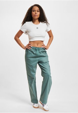 Donna con capelli ricci in piedi con le mani sui fianchi, indossa una t-shirt corta bianca Adidas, pantaloni turchesi e sneakers bianche con strisce viola.