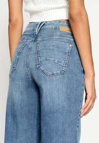 Žena oblečená v světle modrých džínech s vysokým pasem a bílém pleteném crop topu, zobrazená zezadu se jednou rukou uvolněně položenou na boku.