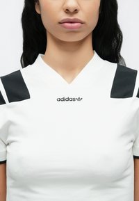 Λευκό αθλητικό μπλουζάκι με μαύρες λεπτομέρειες στους ώμους και σχέδιο σε σχήμα V στο λαιμό. Φέρει το λογότυπο της Adidas σε μαύρο στο κέντρο. Ομαλή υφή.