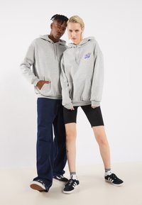 Grå hættetrøjer med kængurulommer, broderet detalje, parret med mørke jeans og sorte cykelshorts. Sneakers med kontrasterende striber.