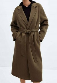 Manteau vert olive ceinturé avec col à revers, manches longues et texture douce, offrant une coupe décontractée et deux poches latérales.
