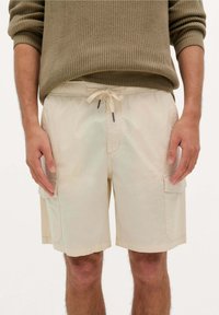 Shorts cargo beige avec une taille à cordon et des poches latérales. Le tissu semble léger avec une texture douce et une coupe décontractée.