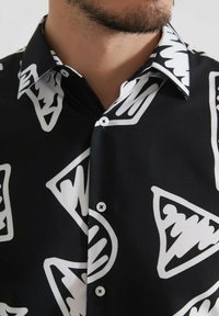 Chemise noire avec des motifs géométriques blancs, un col pointu et une fermeture à boutons sur le devant, fabriquée en tissu lisse avec une texture douce.