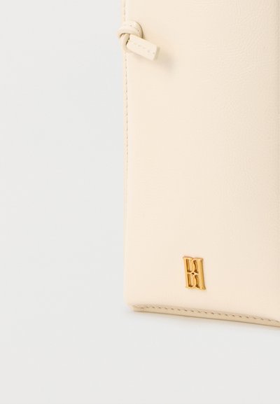 Πορτοφόλι από δέρμα σε κρεμ αποχρώσεις με μικρό χρυσό μεταλλικό λογότυπο και ραμμένο χείλος σε ανοιχτό γκρι φόντο.