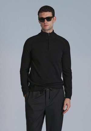 Pull noir à col zippé en tissu doux, avec des manches longues et une coupe ajustée. Assorti à un pantalon sombre à cordon de serrage.