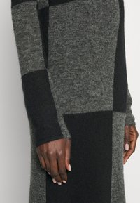 Stickad tröja med växlande mörkgrå, ljusgrå och svarta lappar; mjuk textur; åtsittande mudd; hand som vilar på sömmen.