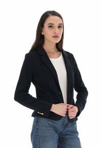 Blazer navy in morbido tessuto, con una vestibilità slim, rever a pattina, due tasche frontali e dettagli in bottone sulle maniche. Indossato sopra una maglia bianca.