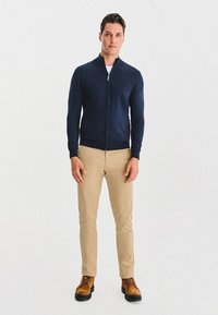 Granatowy sweter na zamek, dopasowany krój, ściągacze na rękawach, połączony z beżowymi spodniami wąskiego kroju i brązowymi sznurowanymi butami na neutralnym tle.