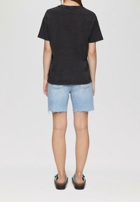 Osoba stojící zády, oblečená v černém tričku, světle modrých džínech s roztrhanými okraji a modrých sandálech na skládání.