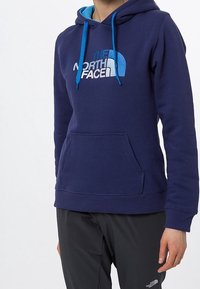 Granatowy hoodie z kieszenią z przodu, kapturem ze sznurkiem i kontrastowymi jasnoniebieskimi akcentami. Ma logo "The North Face" w białym i niebieskim kolorze.