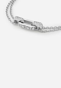 Collana in argento con un link centrale decorato con cristalli trasparenti e un design borchiato, marchiata con "GUESS" su metallo lucido.