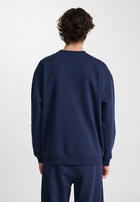 Person med kort lockigt hår som bär en enkel marinblå sweatshirt och matchande byxor, stående med ryggen mot en enfärgad bakgrund.