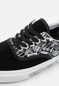 Svarta mockaskor med ett vitt grafiskt mönster, svarta snörningar och vit gummisul med märkesdetalj. Detaljer inkluderar kontrasterande sömmar.