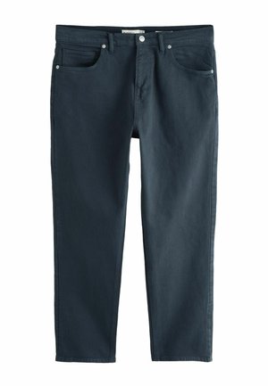 Mann trägt dunkelblaue Jeans, olivgrünes Hemd, schwarze Socken und beige Wildleder-Slipper mit Schnallenriemen, steht vor einer beigen Wand.