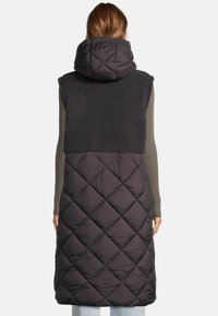 Femme portant un long gilet matelassé noir avec une capuche et un dos en polaire, associé à un haut marron à manches longues et un jean bleu clair.