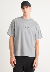 Grå t-skjorte med kort erme og rund hals, med "Calvin Klein" trykket i svart over brystet; kombinert med svarte bukser.