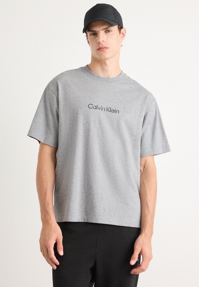 Grå t-skjorte med kort erme og rund hals, med "Calvin Klein" trykket i svart over brystet; kombinert med svarte bukser.