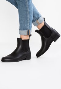 Botas de tobillo negras de goma suave con paneles laterales elásticos, punta redonda y un tacón bajo, combinadas con jeans azules enrollados.